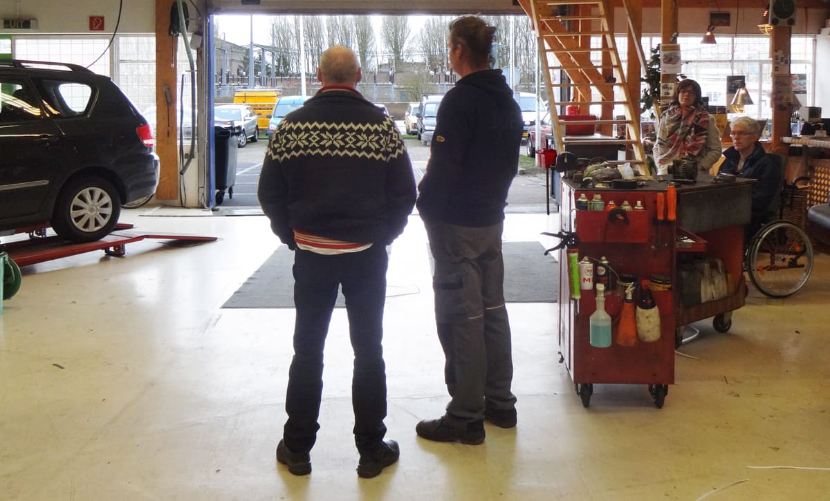 Twee mensen staan in de werkplaats bij een gereedschapswagen te overleggen, met een auto op de hefbrug en de deur open naar het erf.
