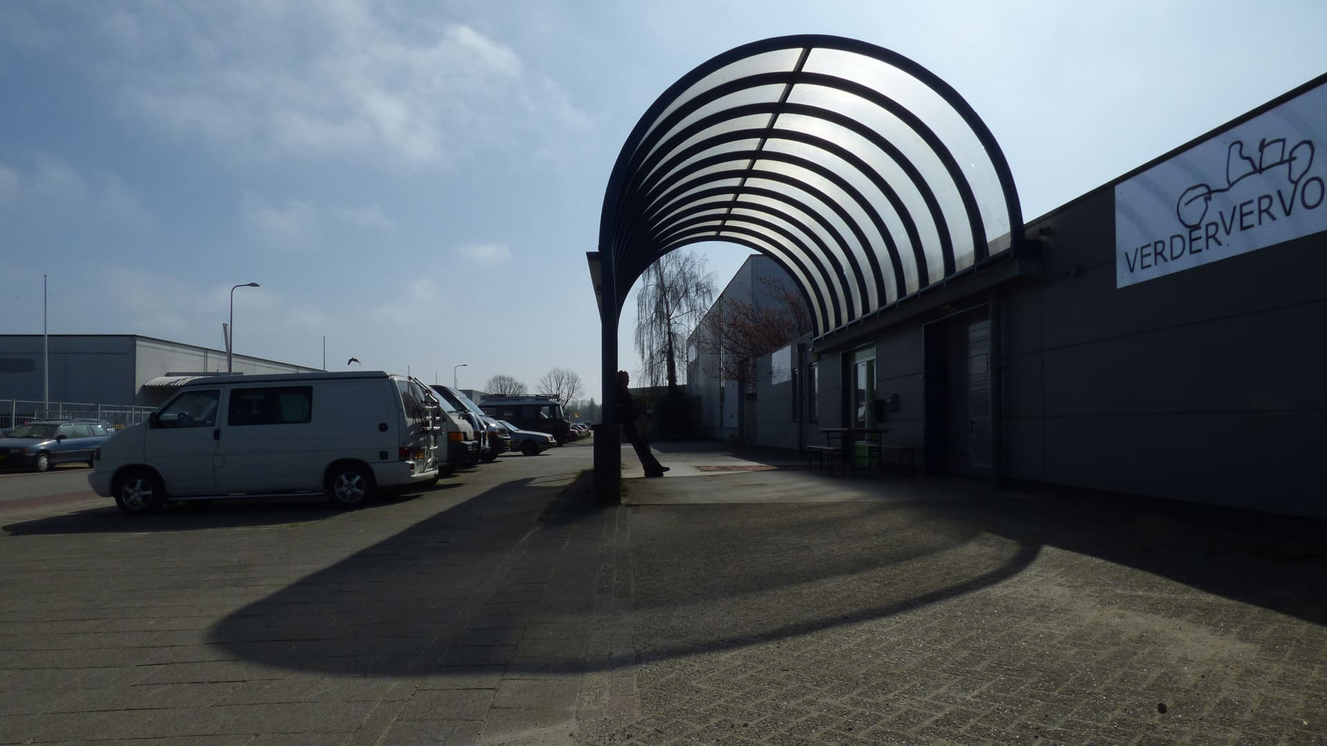 De ingang van Verdervervoer: een gebogen overkapping van staal en lichtdoorlatende platen naast het grijze pand met het bord Verdervervoer.