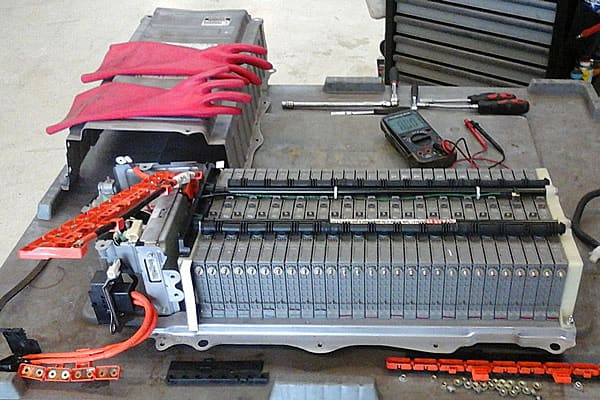 Een geopend accupakket van een hybride auto op de werkbank, met de cellen op een rij, een multimeter en rode werkhandschoenen ernaast.